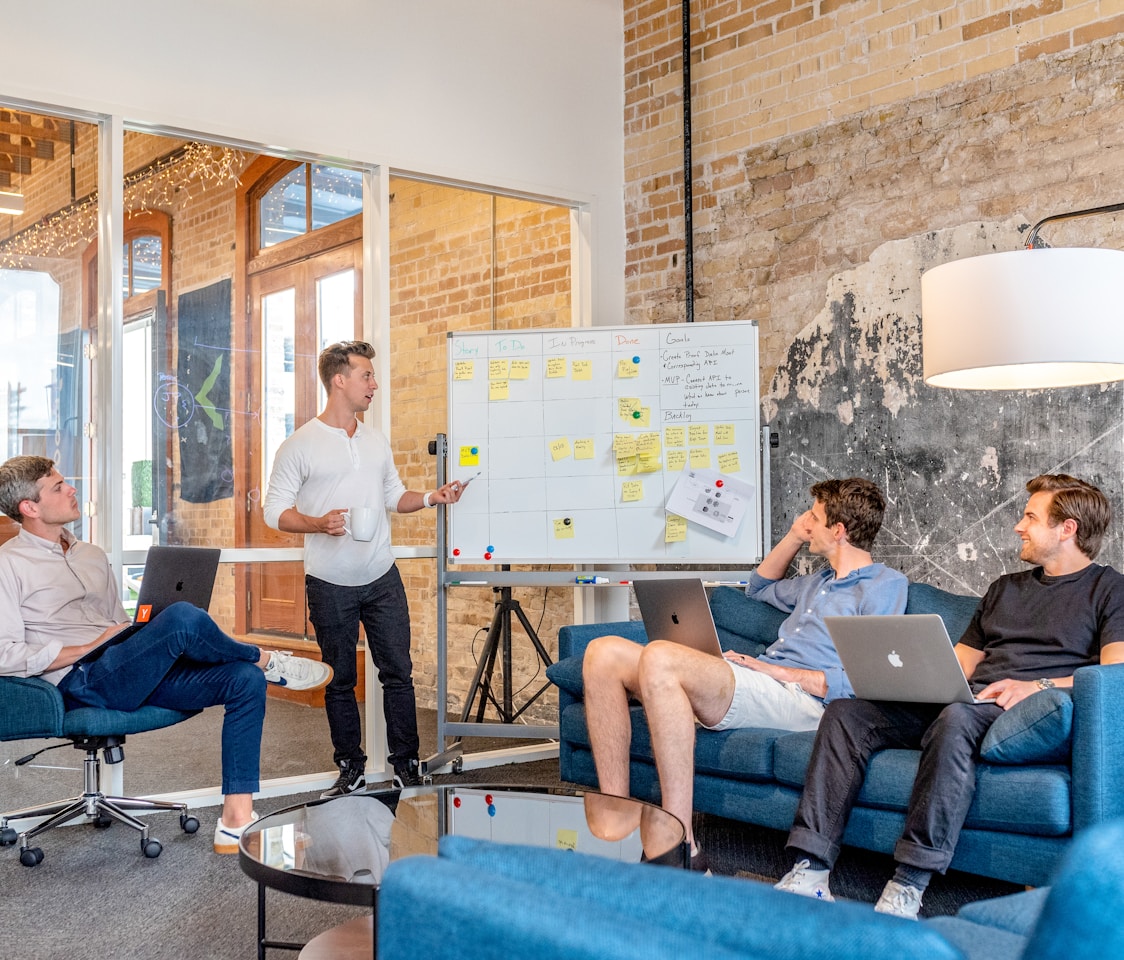 three men sitting while using laptops and watching man beside whiteboard